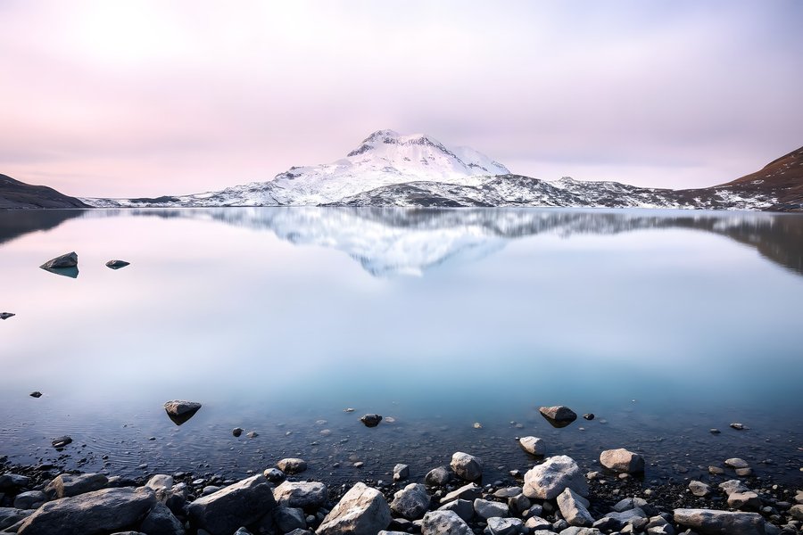A still mountain lake mirroring a snow-capped peak under a pastel pink and violet dawn sky, rocky empty shore, mountain fine art print.
